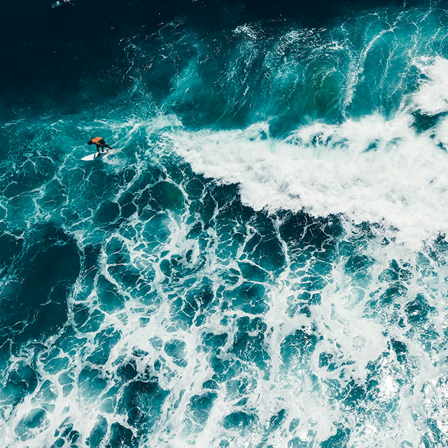 Meer. Das Wasser durch eine Welle aufgeschäumt. Ein Surfer der auf der auslaufenden Welle reitet.