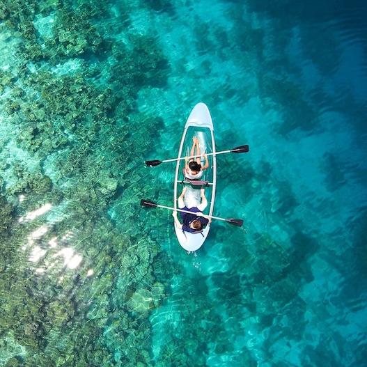 Zwei Personen, die in einem Kajak im Meer paddeln. Das Wasser ist so klar, dass man den Meeresgrund und die darauf wachsenden Korallen sehen kann. Das Foto ist direkt über dem Kajak, aus der Perspektive einer Drone aufgenommen.